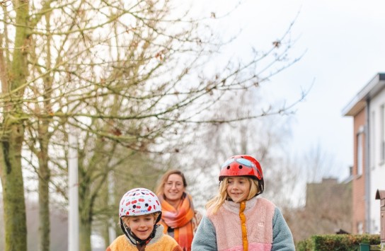 Twee kinderen met helmen rijden op step, gevolgd door een vrouw op een trottoir in een woonwijk. (vergroot foto)