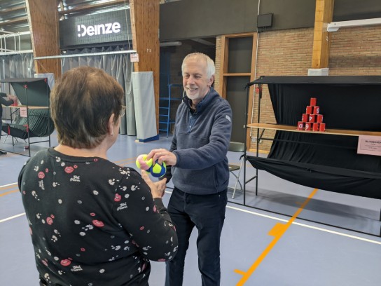 Twee mensen in een sporthal spelen met zachte ballen, op de achtergrond een stapel rode blikken. (vergroot foto)