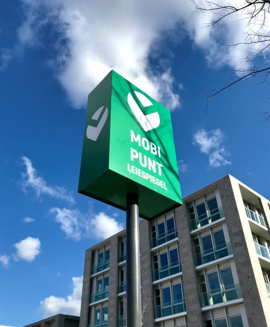 Groen bord met tekst "Mobi Punt Leiespiegel" tegen een blauwe lucht en modern gebouw op de achtergrond. (vergroot foto)