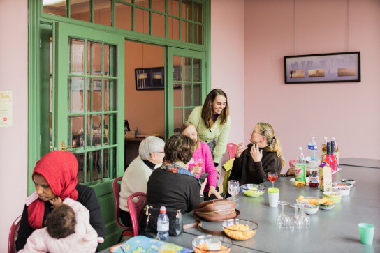 Groep vrouwen zit samen aan tafel met hapjes en drankjes, in een gezellige kamer met groene deuren. (vergroot foto)