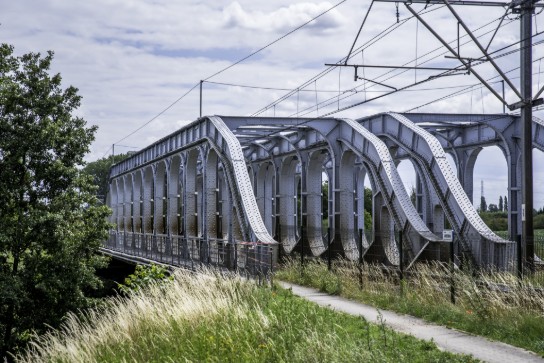 Stalen spoorbrug met boogvormige steunbalken over een rivier, omgeven door gras en bomen. (vergroot foto)