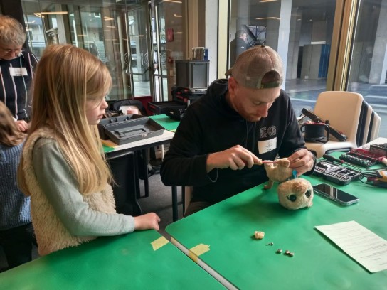 Man repareert een knuffel terwijl een meisje toekijkt aan een tafel met gereedschap. (vergroot foto)