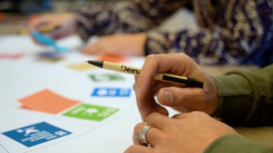 Hand met pen boven een tafel met kleurrijke kaarten en papieren, mensen op de achtergrond. (vergroot foto)