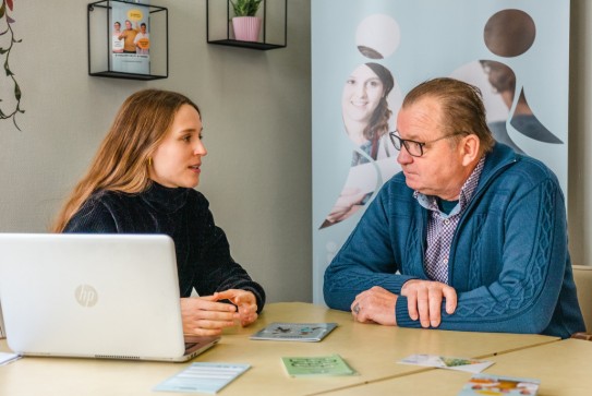 Twee mensen in gesprek aan tafel met laptop en folders, in een kantooromgeving. (vergroot foto)