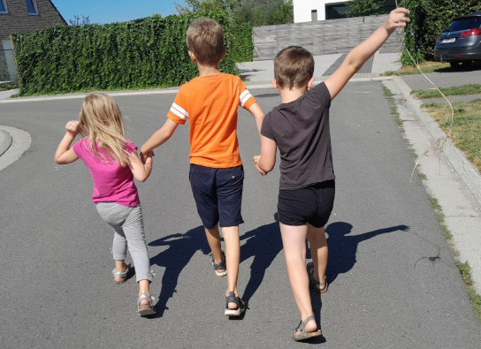Drie kinderen lopen hand in hand over een straat op een zonnige dag, één zwaait met een tak. (vergroot foto)
