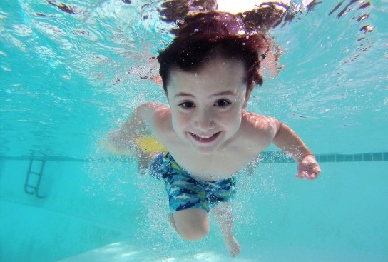 Jongentje zwemt onder water in een zwembad met blauwe zwemshorts en kijkt lachend naar de camera. (vergroot foto)