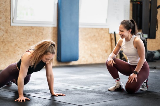 Twee vrouwen in sportkleding trainen samen in een sportschool, één doet push-ups, de ander moedigt aan. (vergroot foto)