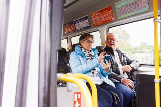 Twee oudere mensen zitten samen in een bus, een vrouw toont iets op haar smartphone aan een man. (vergroot foto)