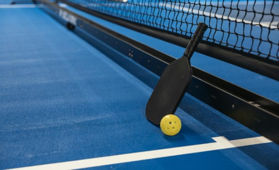 Zwarte pickleball paddle en gele bal liggen tegen het net op een blauwe indoor pickleballbaan. (vergroot foto)
