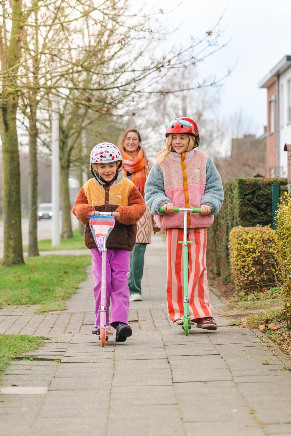 Twee kinderen met helmen rijden op steppen, gevolgd door een vrouw op een trottoir in een woonwijk.