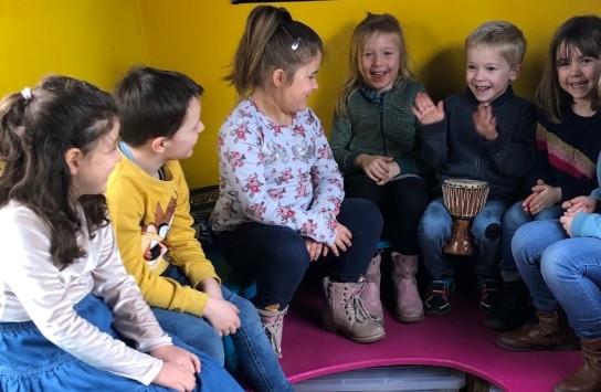 Stedenbandhuisje: Kinderen zitten in een kring en kijken lachend naar poppen en speelgoed op een tafel met een geel kleed. (vergroot foto)