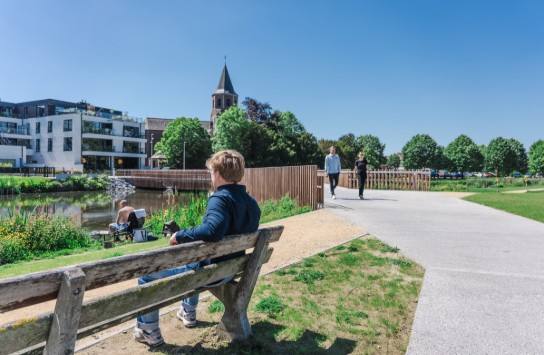 Man zit op bankje aan de Leie, op de achtergrond kerk, wandelend paar en moderne gebouwen onder blauwe lucht. (vergroot foto)