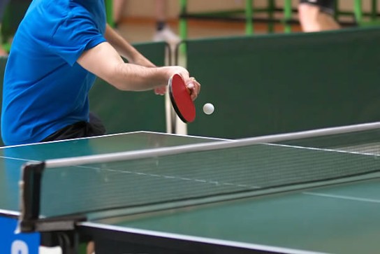 Persoon in blauw t-shirt speelt tafeltennis en slaat de bal met een rode bat. (vergroot foto)