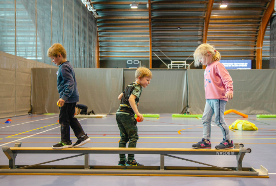 Drie kinderen balanceren op een smalle houten balk in een gymzaal met sportvloeren en hoge wanden. (vergroot foto)