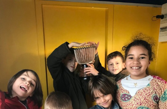 Stedenbandhuisje: Zes kinderen in kleurrijke kleding poseren vrolijk voor een gele muur, één houdt een trommel vast. (vergroot foto)
