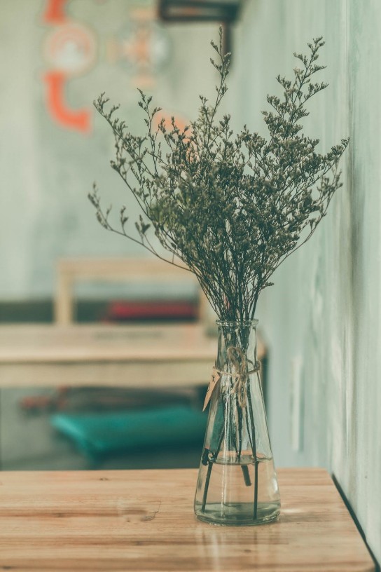 Vaas met takken en kleine bloemen op een houten tafel tegen een lichtblauwe muur. (vergroot foto)